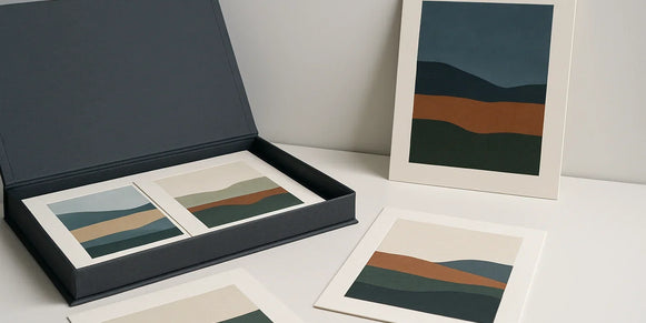 Open charcoal archival portfolio with four loose abstract landscape prints and one slim mounted print on a cool-daylight gallery table