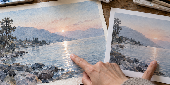 Watercolor original and physical proof compared on a worktable, showing paper-white glints amid pale washes.