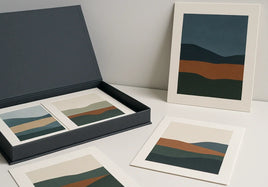 Open charcoal archival portfolio with four loose abstract landscape prints and one slim mounted print on a cool-daylight gallery table
