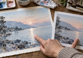 Watercolor original and physical proof compared on a worktable, showing paper-white glints amid pale washes.