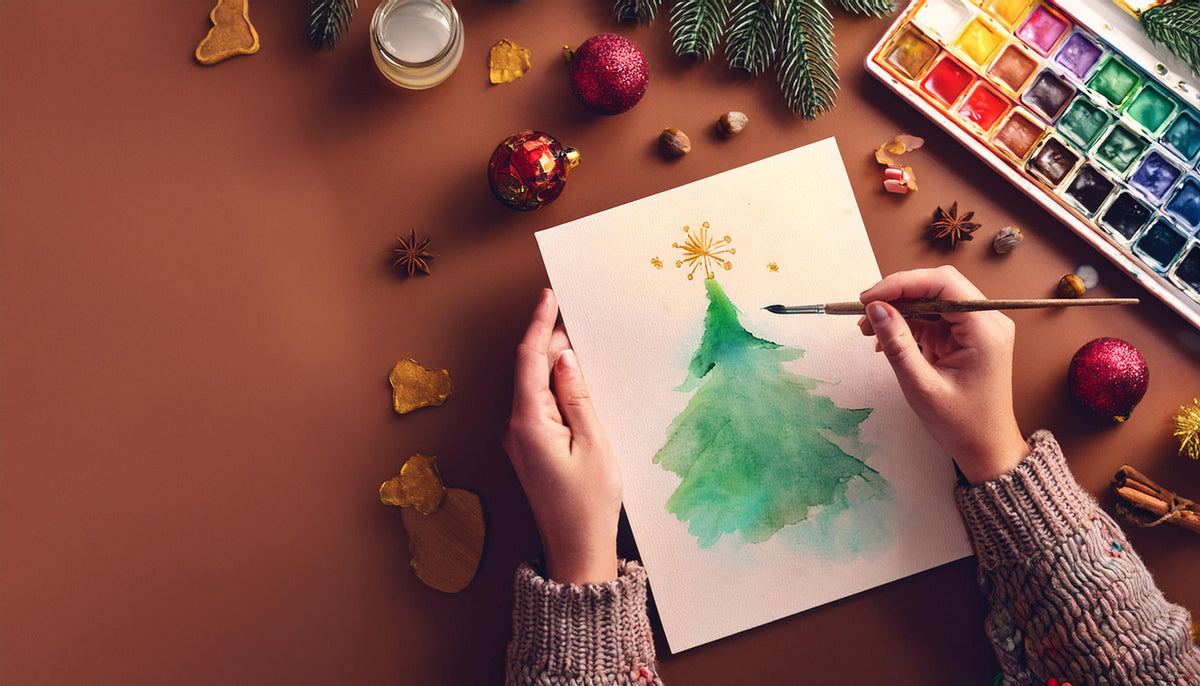 Hands painting a watercolor Christmas tree beside a palette, ornaments, and evergreen branches